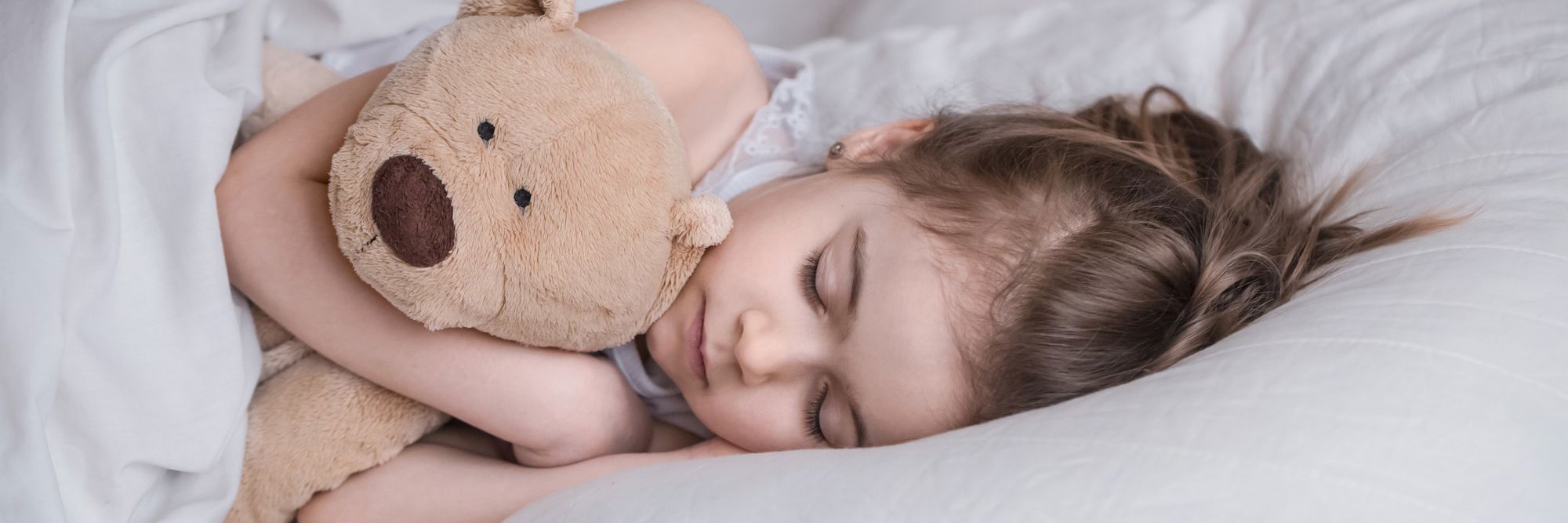 Lille pige ligger i seng med hvidt sengetøj og sover, mens hun krammer en lysebrun bamse