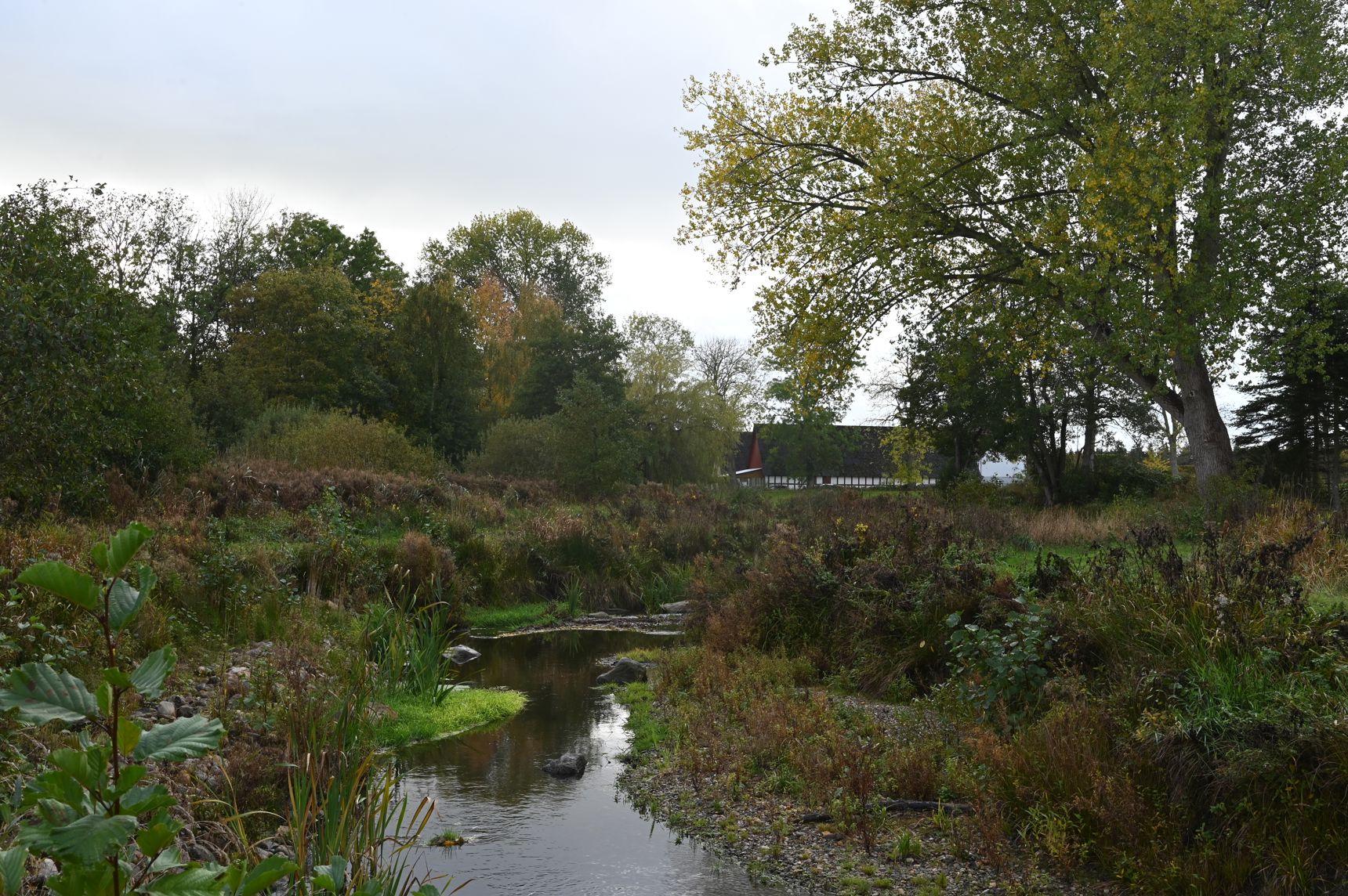 I forgrunden et lavvandet vandløb og i baggrunden en rød/hvid bindingsgsværksgård. Mange træer i kanterne