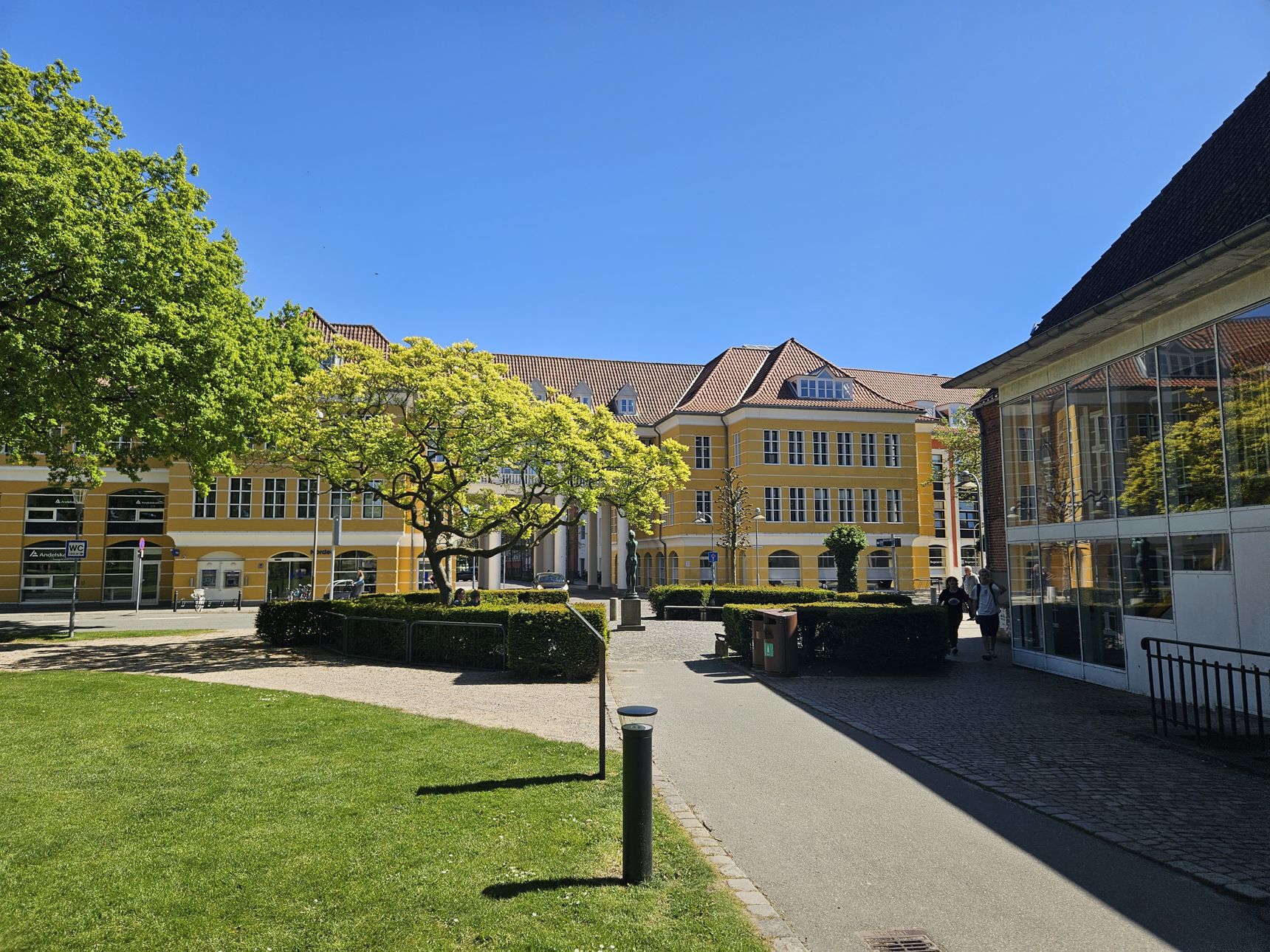 En sti med græs omkring, fører ned til en stor gul bygning med hvide vinduer. Foran bygningen er en plads omkranset af en rund hæk og et stort træ.