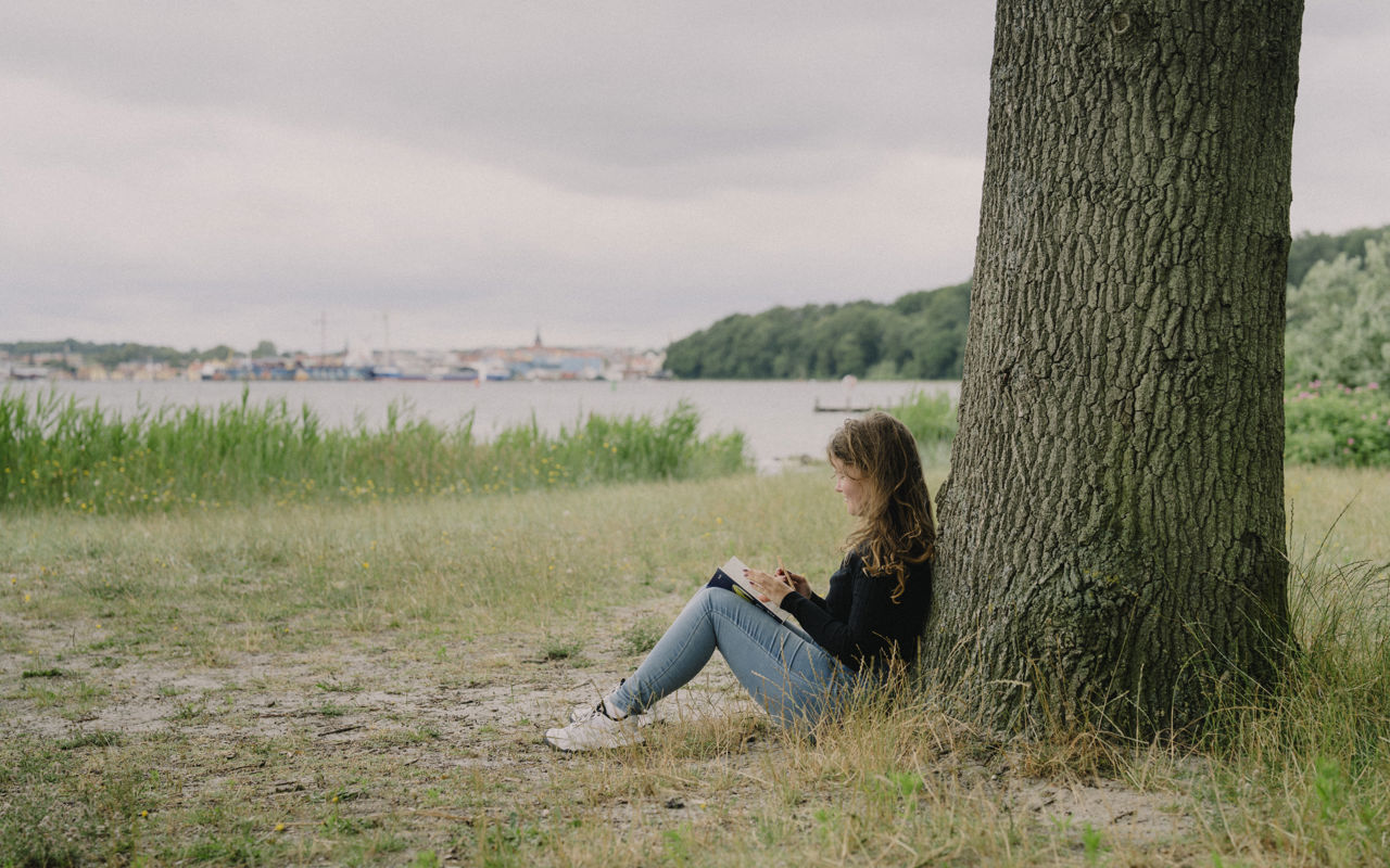 Pige sidder op ad træ og læser bog - i baggrunden Svendborg By set fra vandsiden