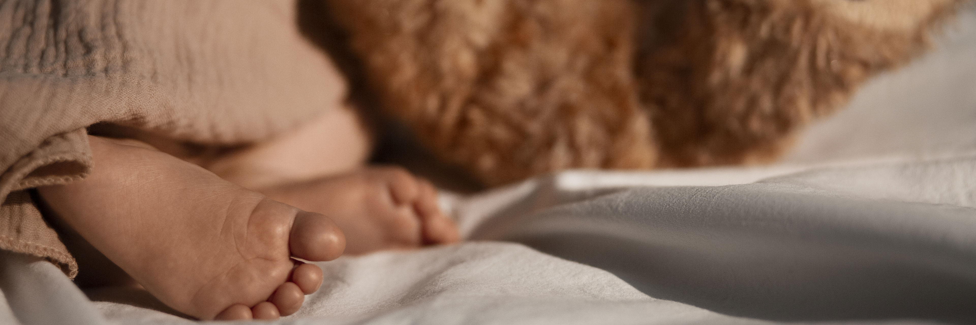 Baby fødder der stikker ud under tæppe. Brun bamses fødder ligger og stritter i vejret ved siden af baby.