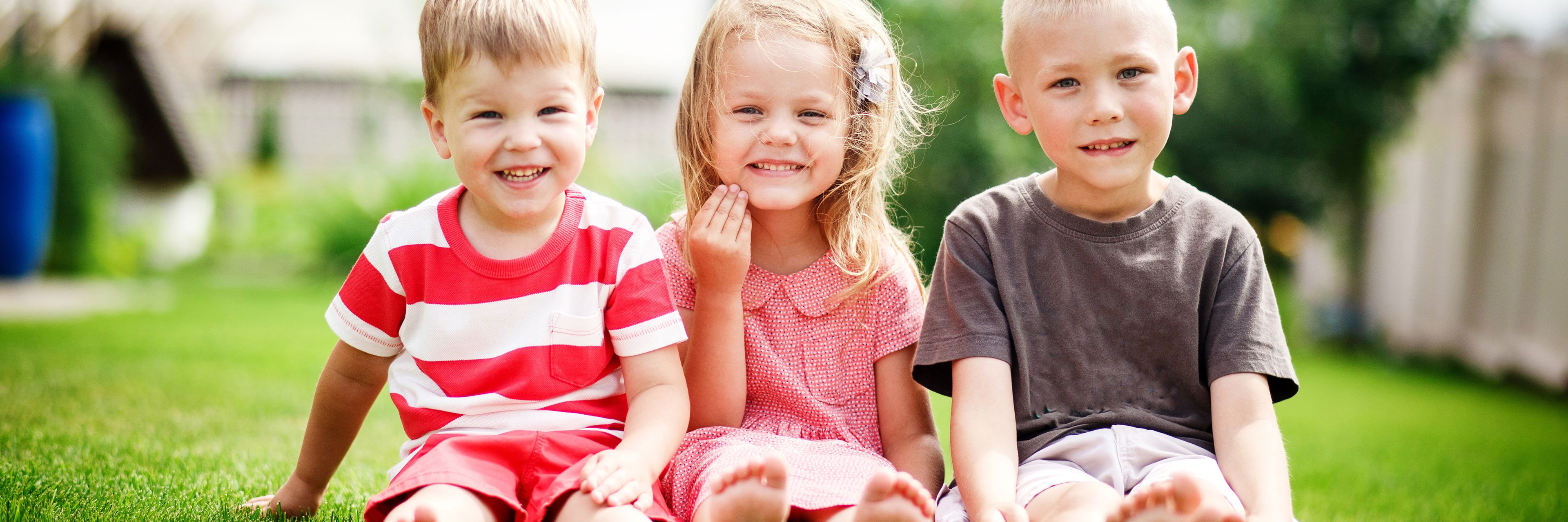 Tre mindre børn sidder på græsset ved siden af hinanden