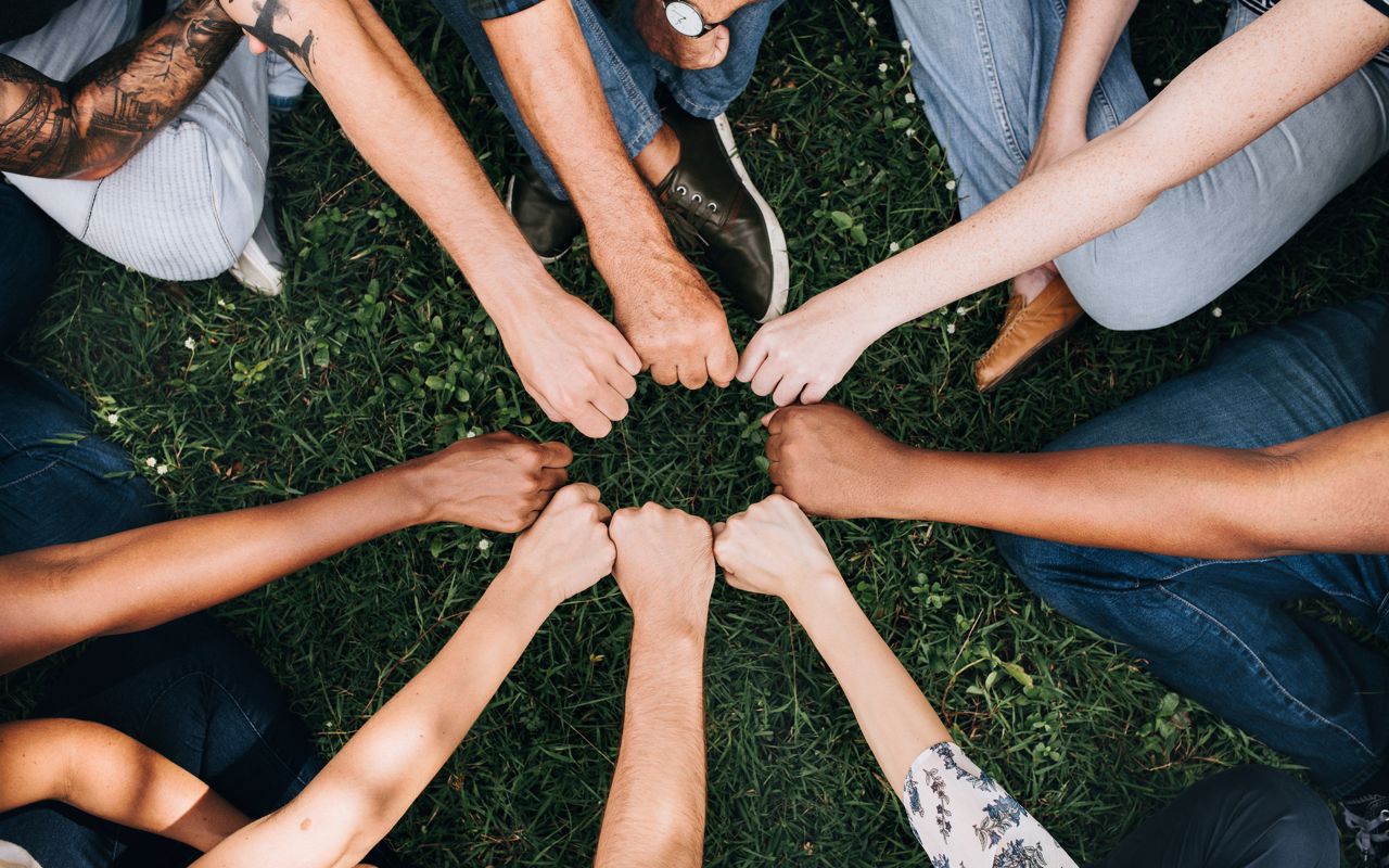 Forskellige mennesker rækker deres knyttede hænder ind mod hinanden, så de danner en cirkel. Billedet signalerer fællesskab og sammenhold.
