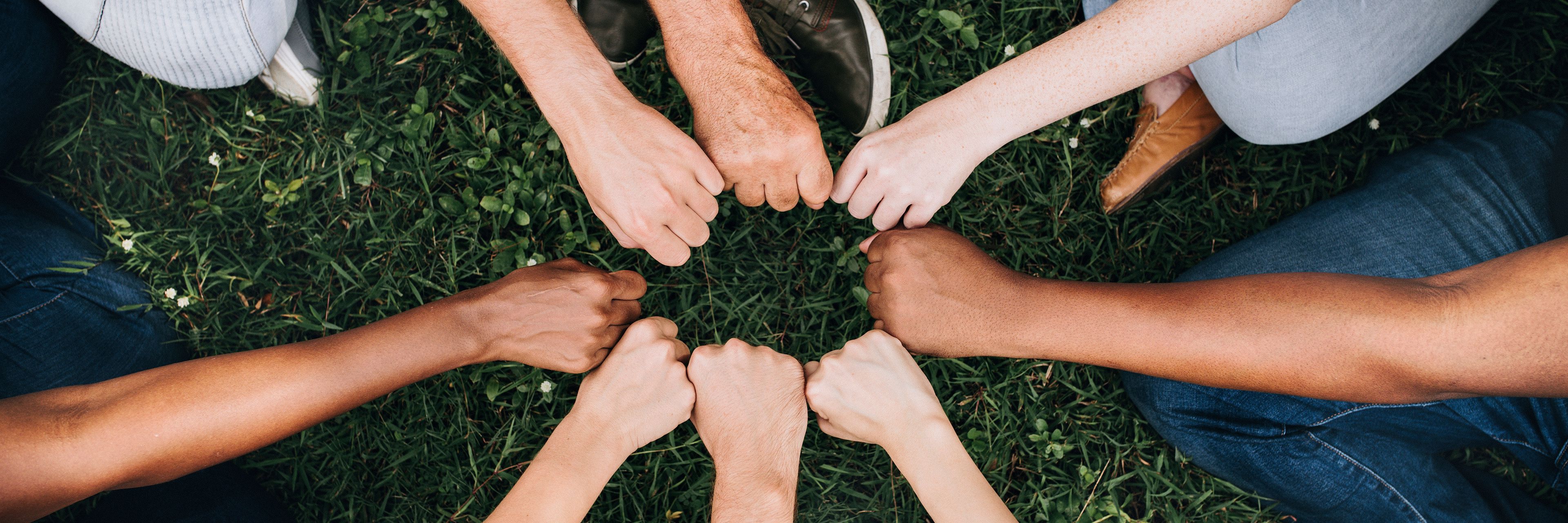 Forskellige mennesker rækker deres knyttede hænder ind mod hinanden, så de danner en cirkel. Billedet signalerer fællesskab og sammenhold.