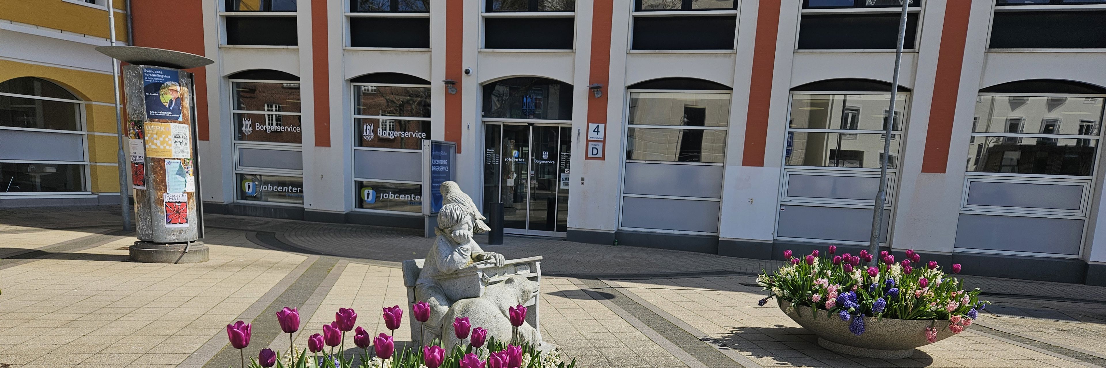 En kumme med forårsblomster foran indgangen til en stor bygning, der er hvid og rød.